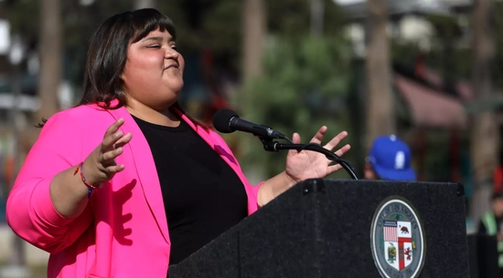 Councilmember Hernandez at the podium with arms outstretched wearing a pink blazer