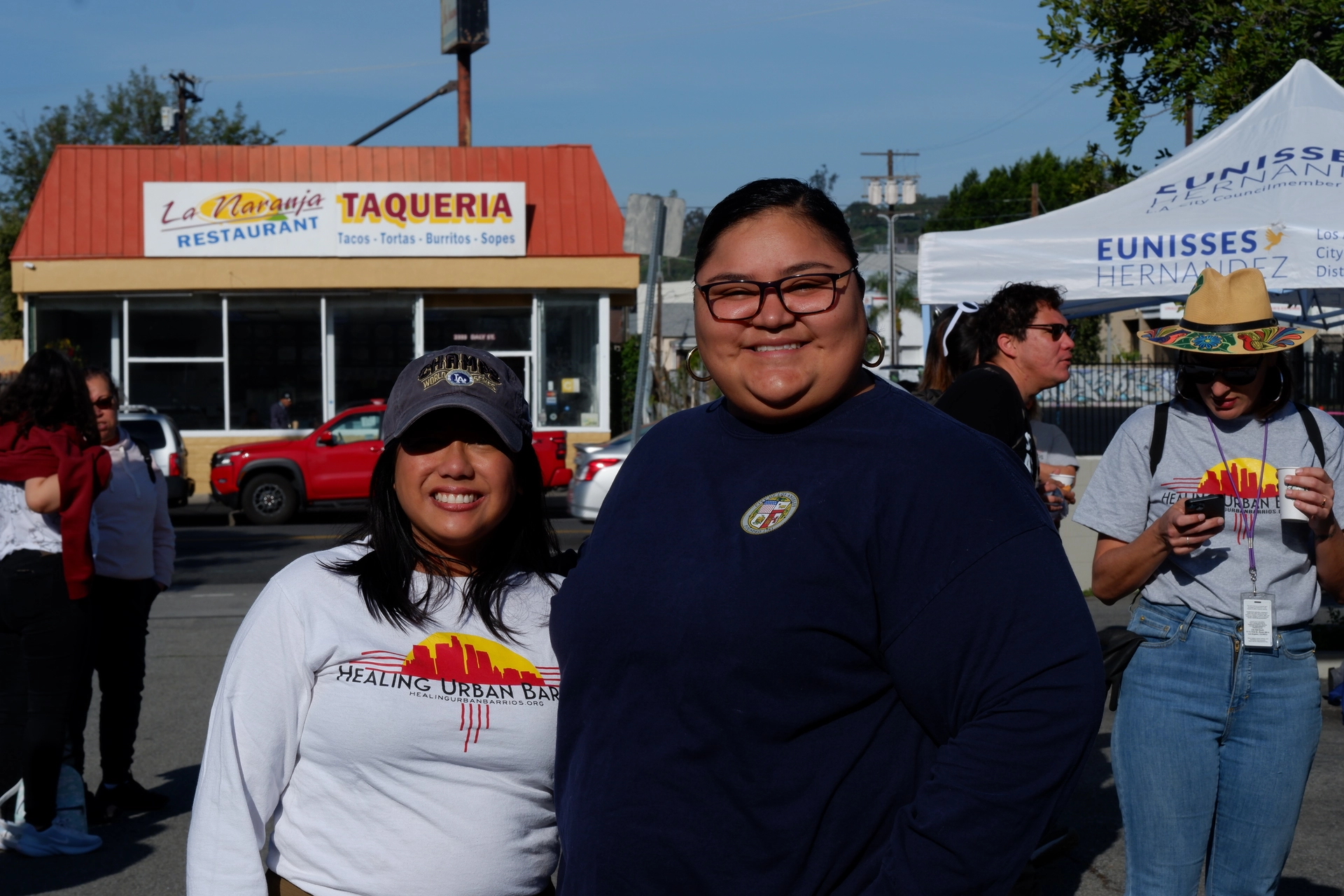 Councilmembers Jurado and Hernandez are smiling in front of a taqueria with others in the background.