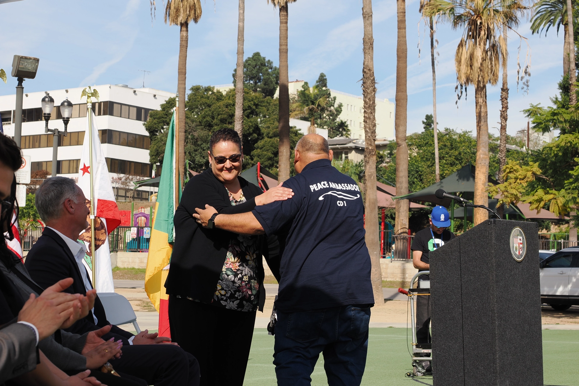 A "Peace Ambassador" greets a woman warmly at an outdoor event. Attendees applaud in a park setting with palm trees and flags.
