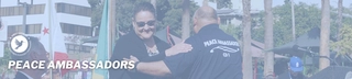 Two people warmly greet at a MacArthur Park press conference with flags and trees; one wears a Peace Ambassadors t-shirt.